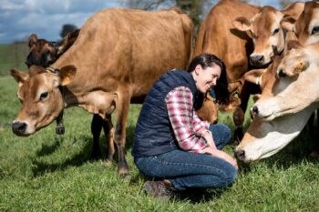 mujer sonriente se agacha junto a las vacas en un prado que la observan curiosas